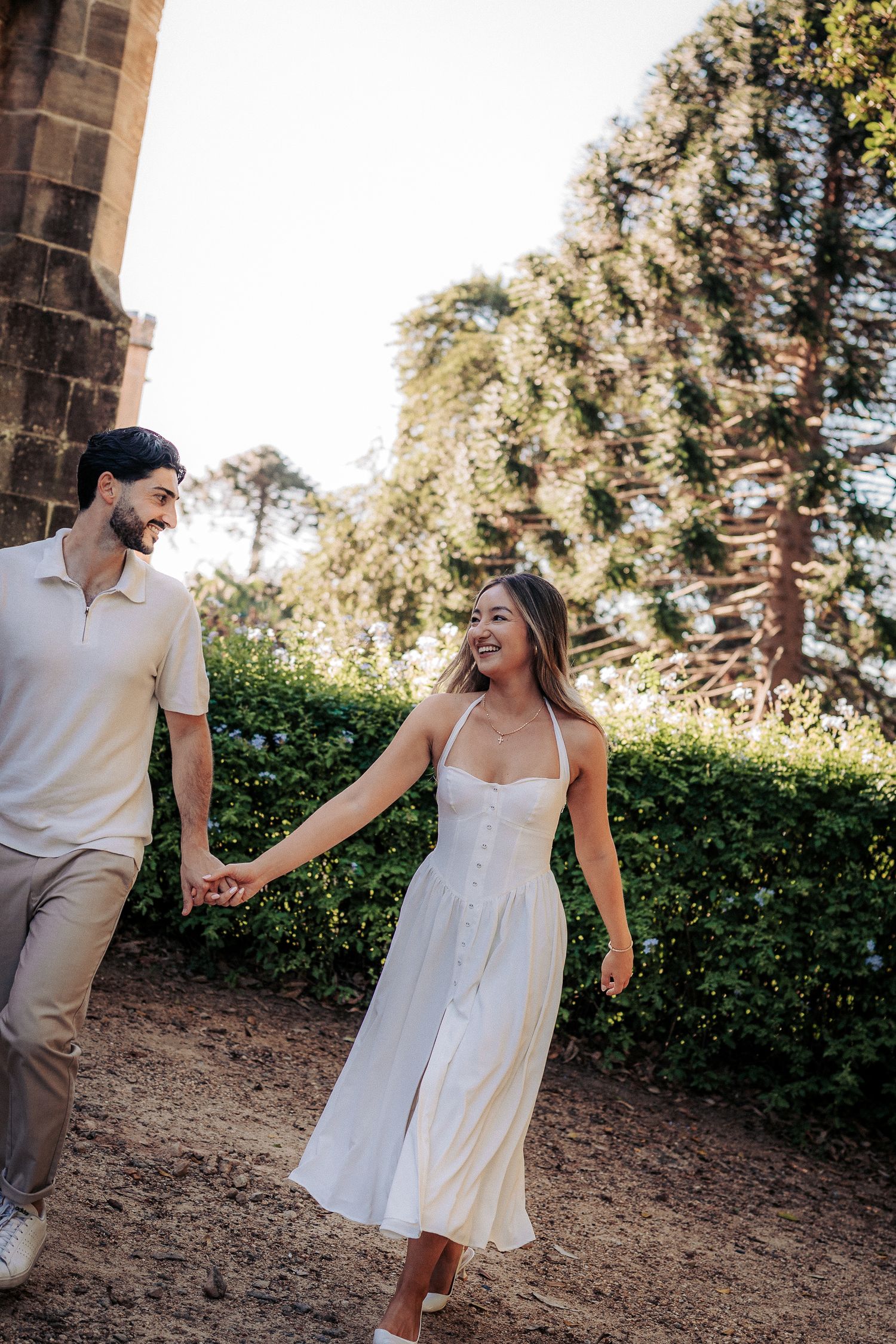 Smiling couple holding hands and walking through a sunlit garden path, woman in white sundress.