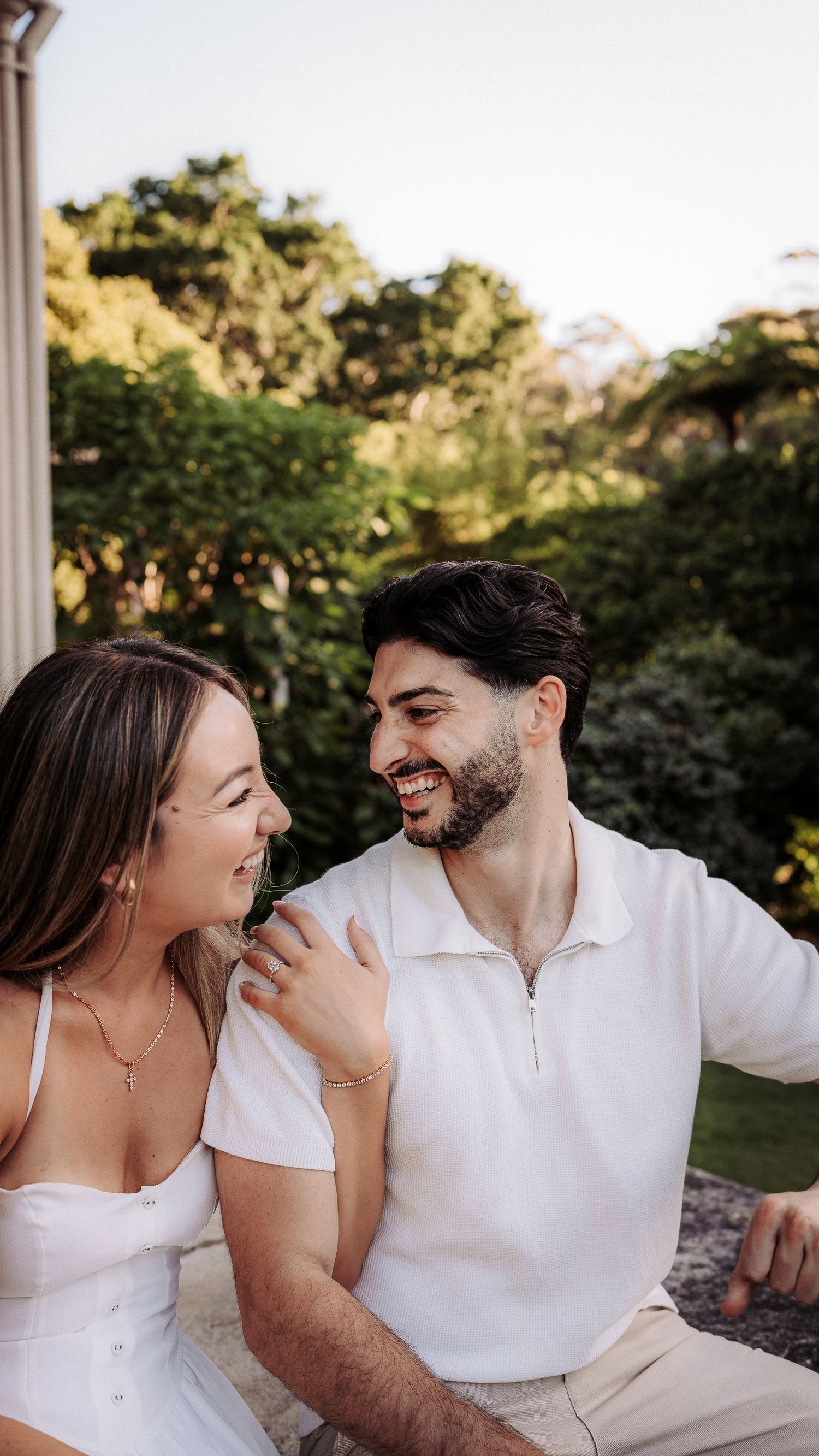 Happy couple laughing together outdoors, woman in white dress touching man's shoulder in garden.