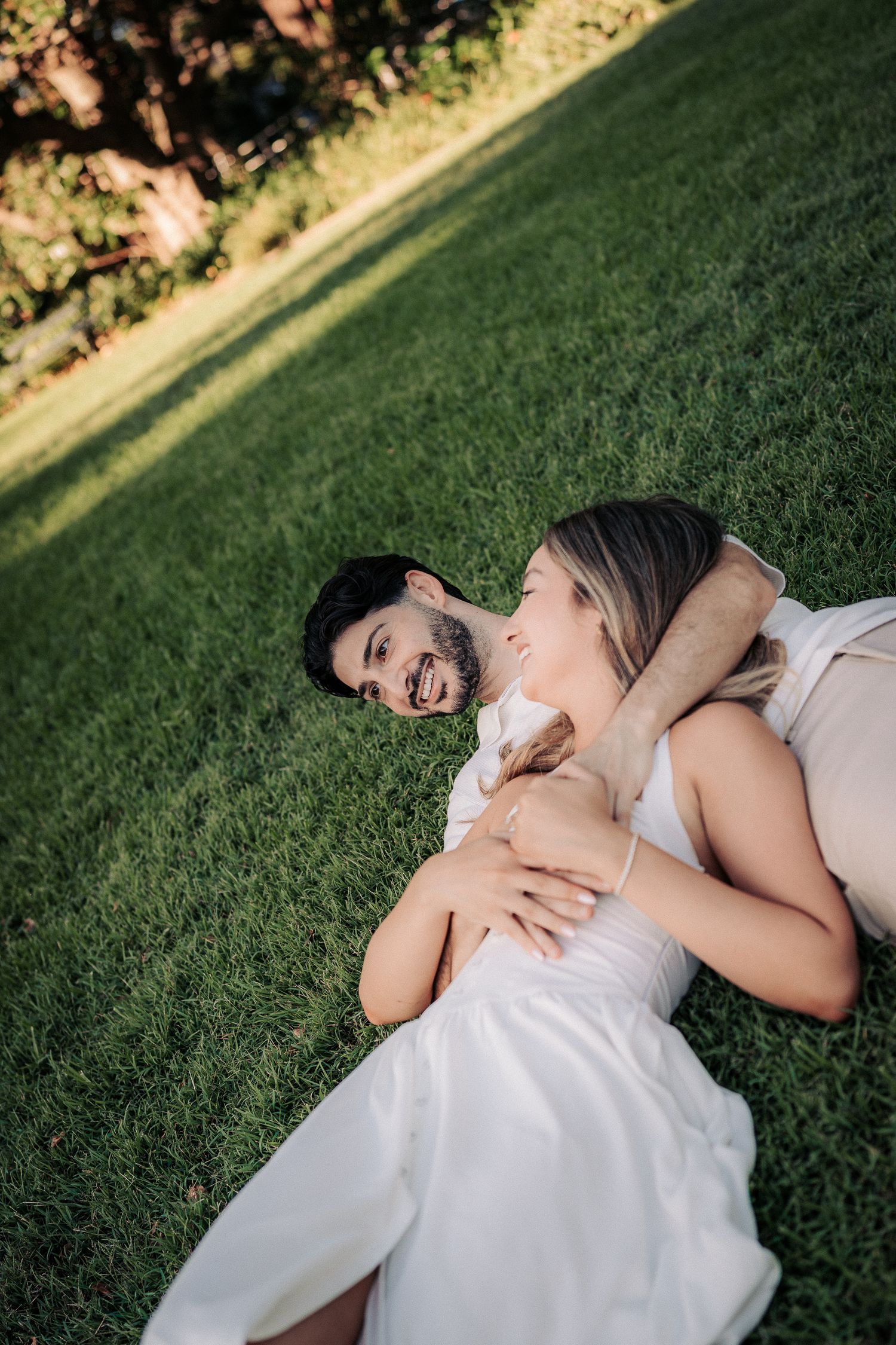 A happy couple lying on green grass, embracing and smiling at each other during an outdoor engagement photo session.