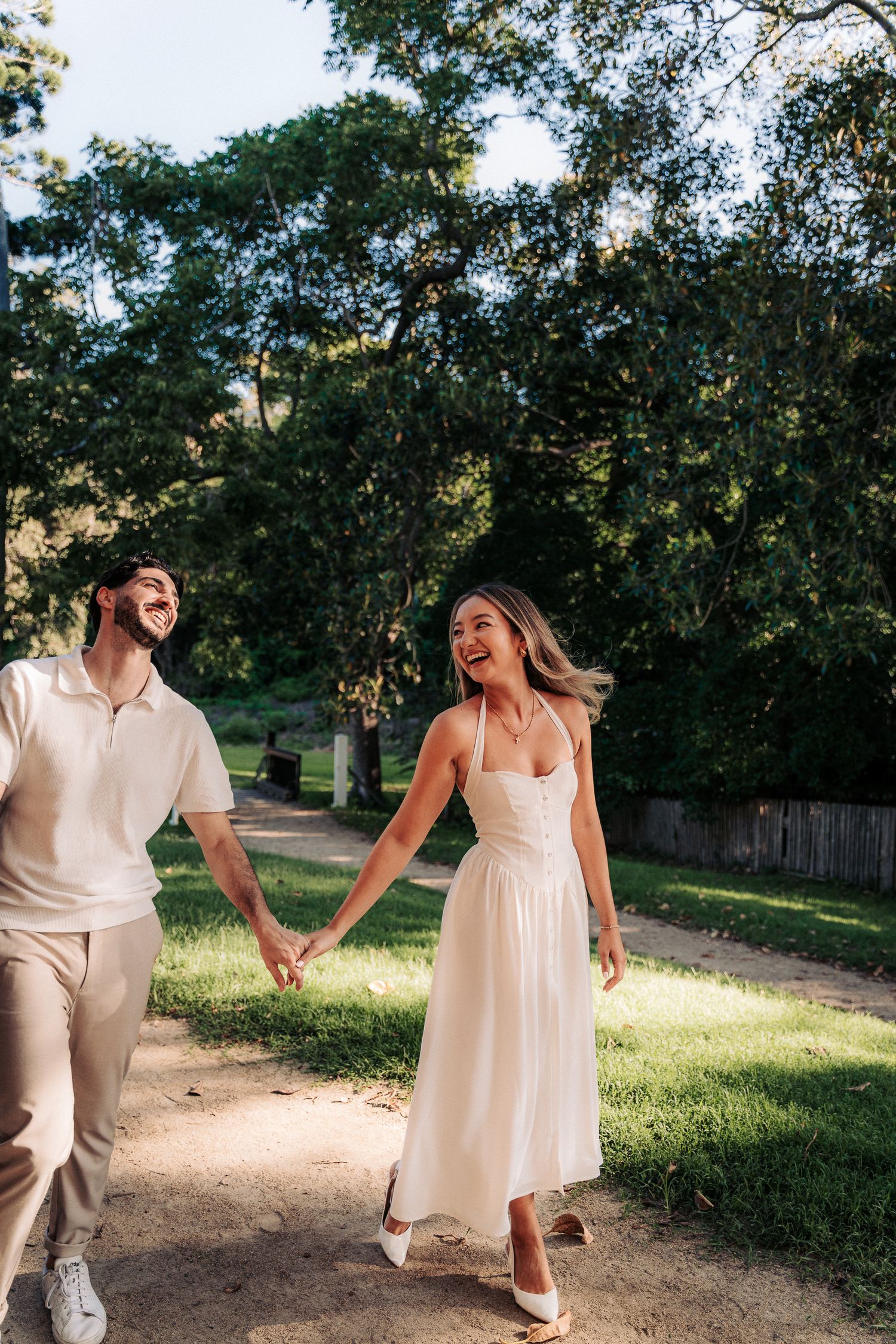 Happy couple holding hands walking on a sunny park path, woman in white midi dress, man in cream polo shirt.