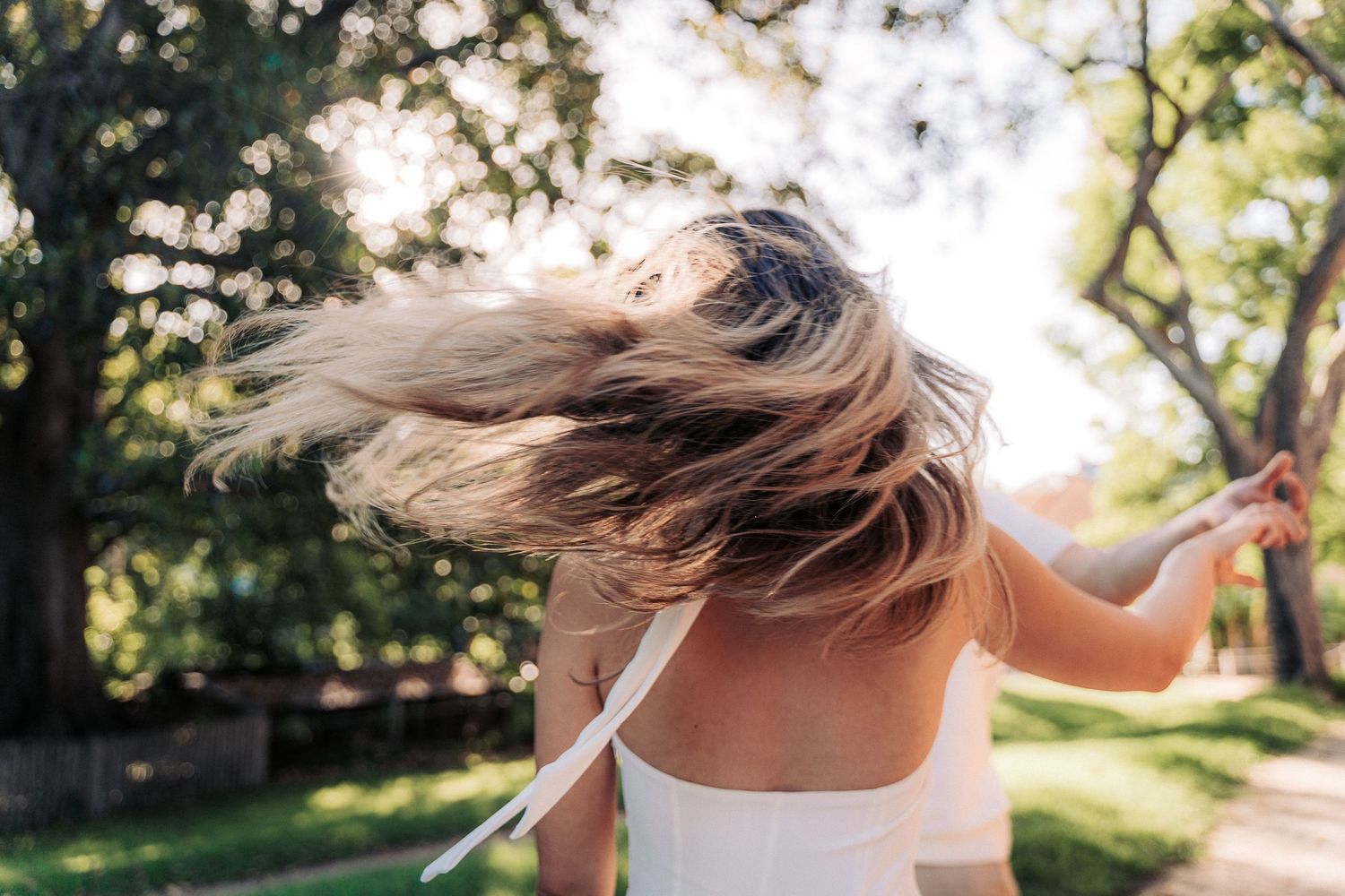A young woman with blonde hair twirling outdoors in a sunny park, wearing a white top, with trees in the background.