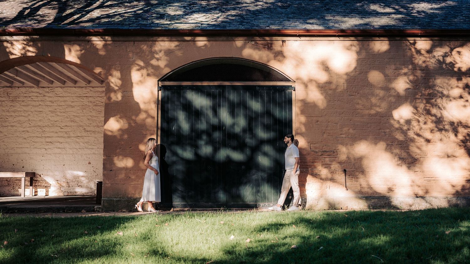 Couple standing by a rustic brick barn with an arched green door, surrounded by dappled sunlight and lush green grass.