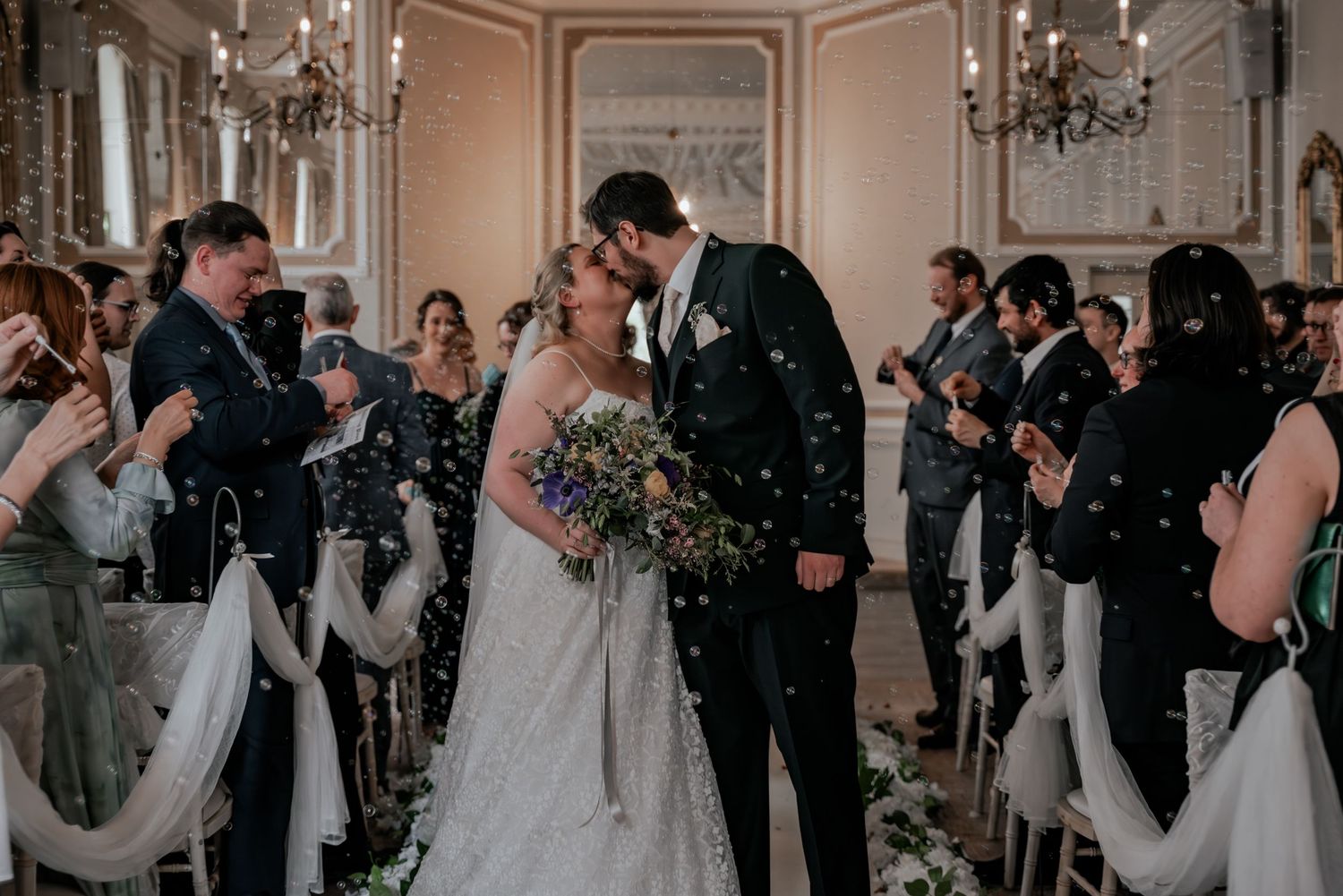 Bride and groom share first kiss at wedding ceremony while guests throw confetti in elegant chandelier ballroom.