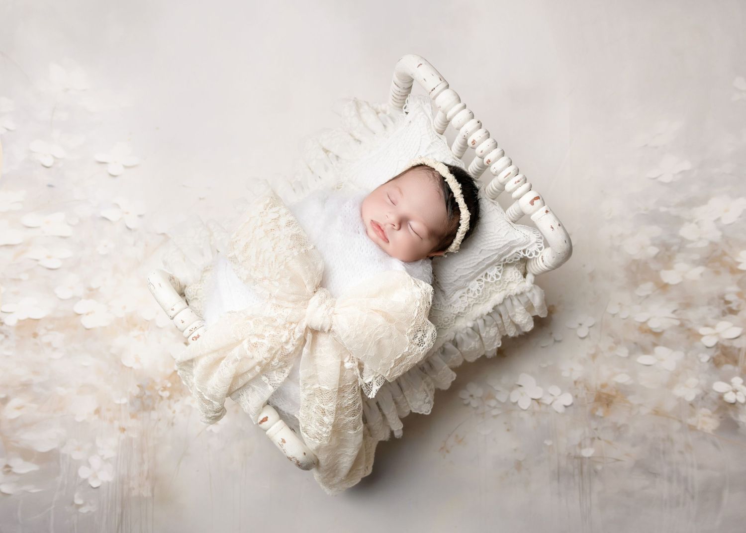 Newborn baby sleeping peacefully in a white lace-draped miniature bed with a delicate floral headband.