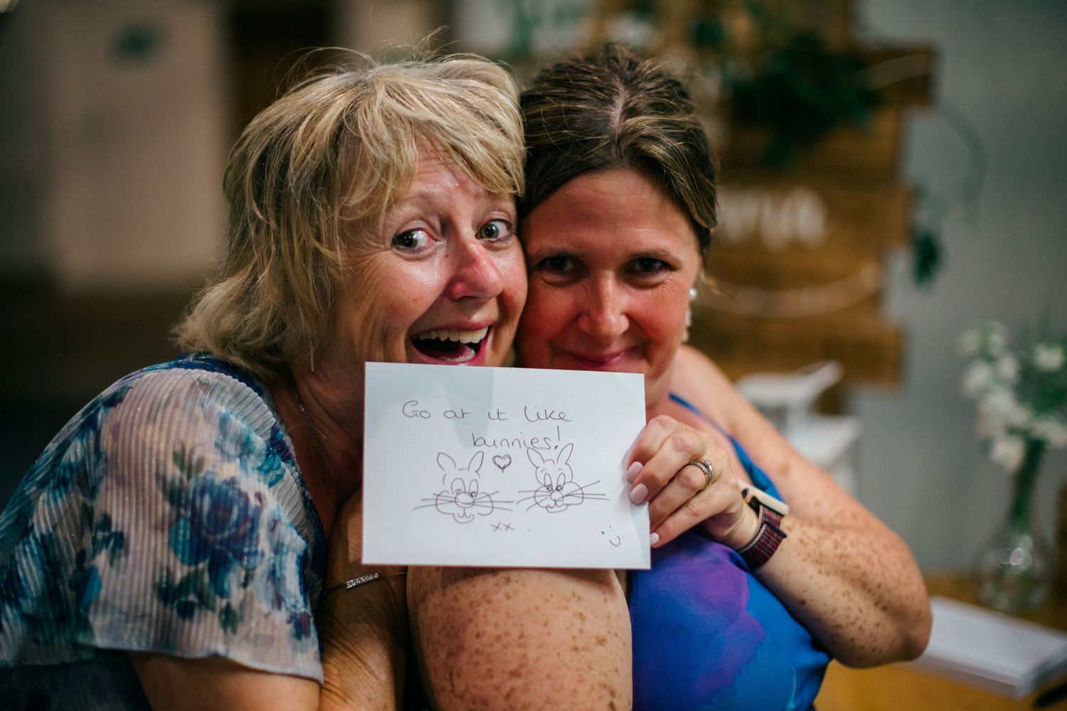 Two women hugging and smiling while holding a handwritten card with a heartfelt message at a celebration.