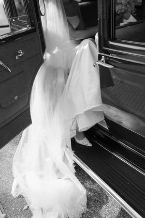 Black and white photo of a bride in a white gown stepping into a vintage car on her wedding day.