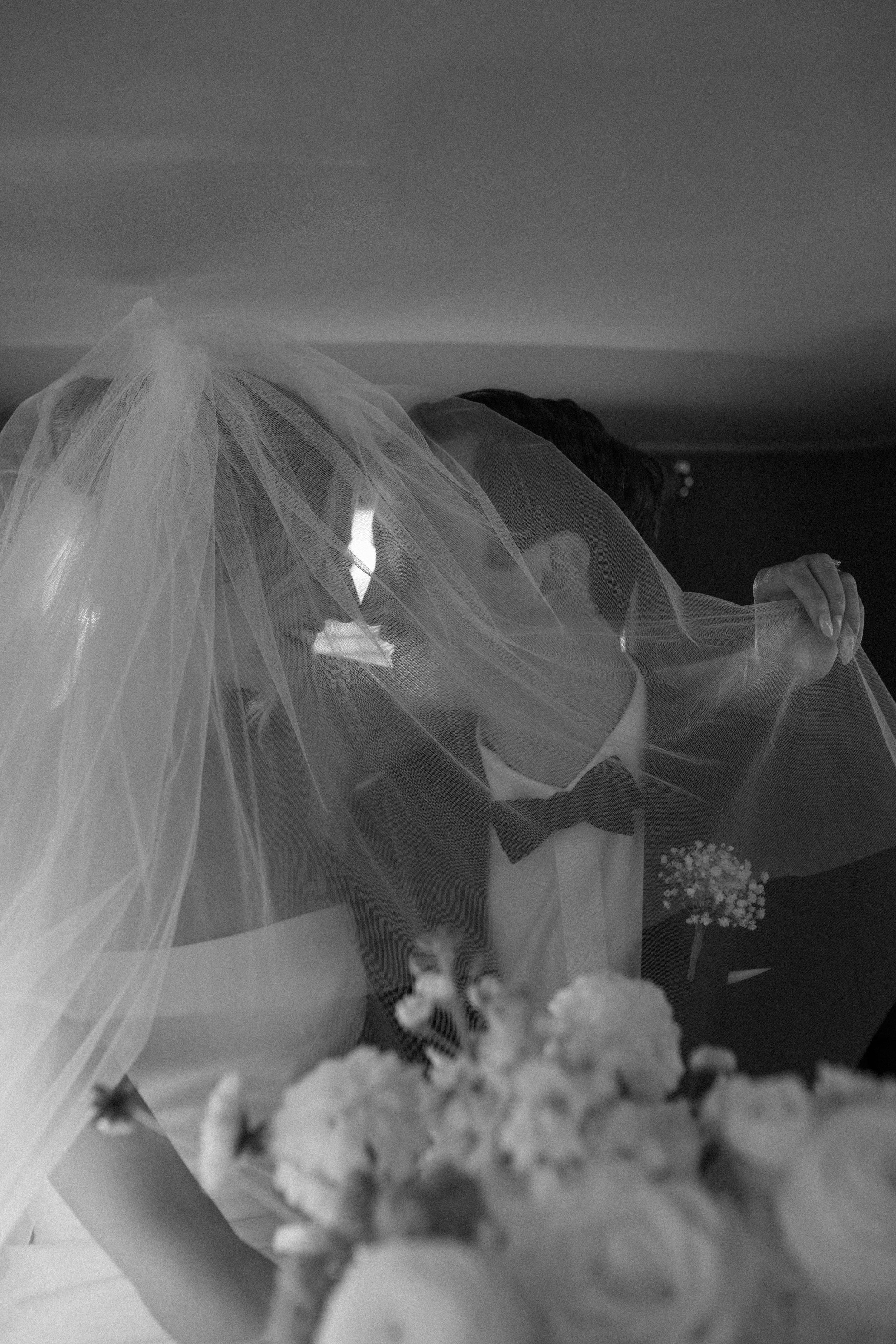 Black and white photo of bride and groom kissing under wedding veil with bouquet in foreground.