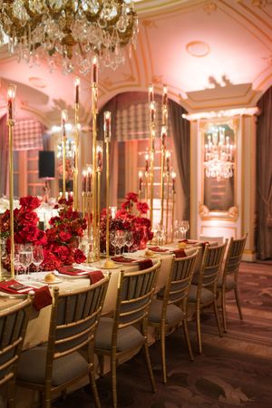 Elegant banquet table with red floral centerpieces, gold candelabras, and crystal chandeliers in a luxurious ballroom.