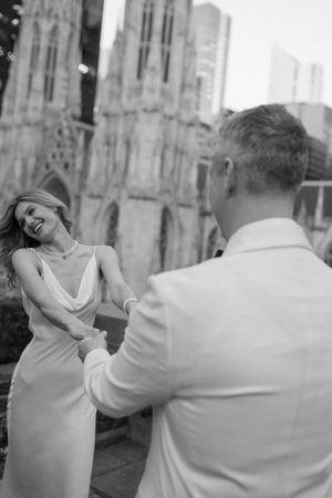 Black and white photo of a smiling bride in a slip dress holding hands with a groom near a Gothic cathedral.