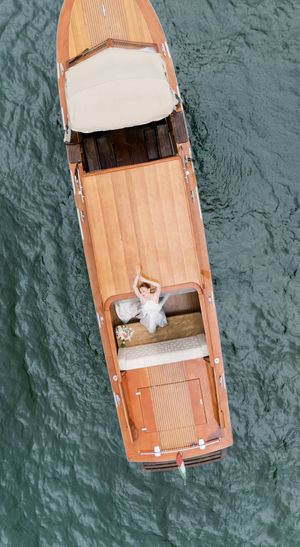 Aerial view of a bride in a white wedding dress lying on a classic wooden luxury boat on turquoise water.