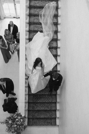 Aerial black and white photo of bride and groom descending elegant staircase, wedding veil flowing, guests watching below.