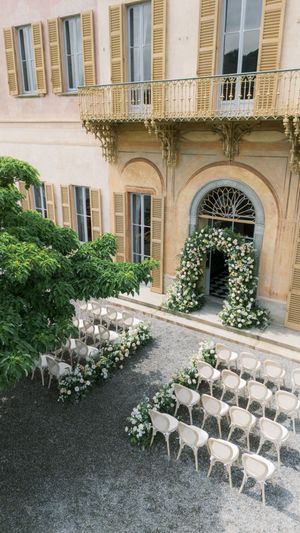 Elegant outdoor wedding ceremony setup with white chairs and floral arch at a classic Italian villa.