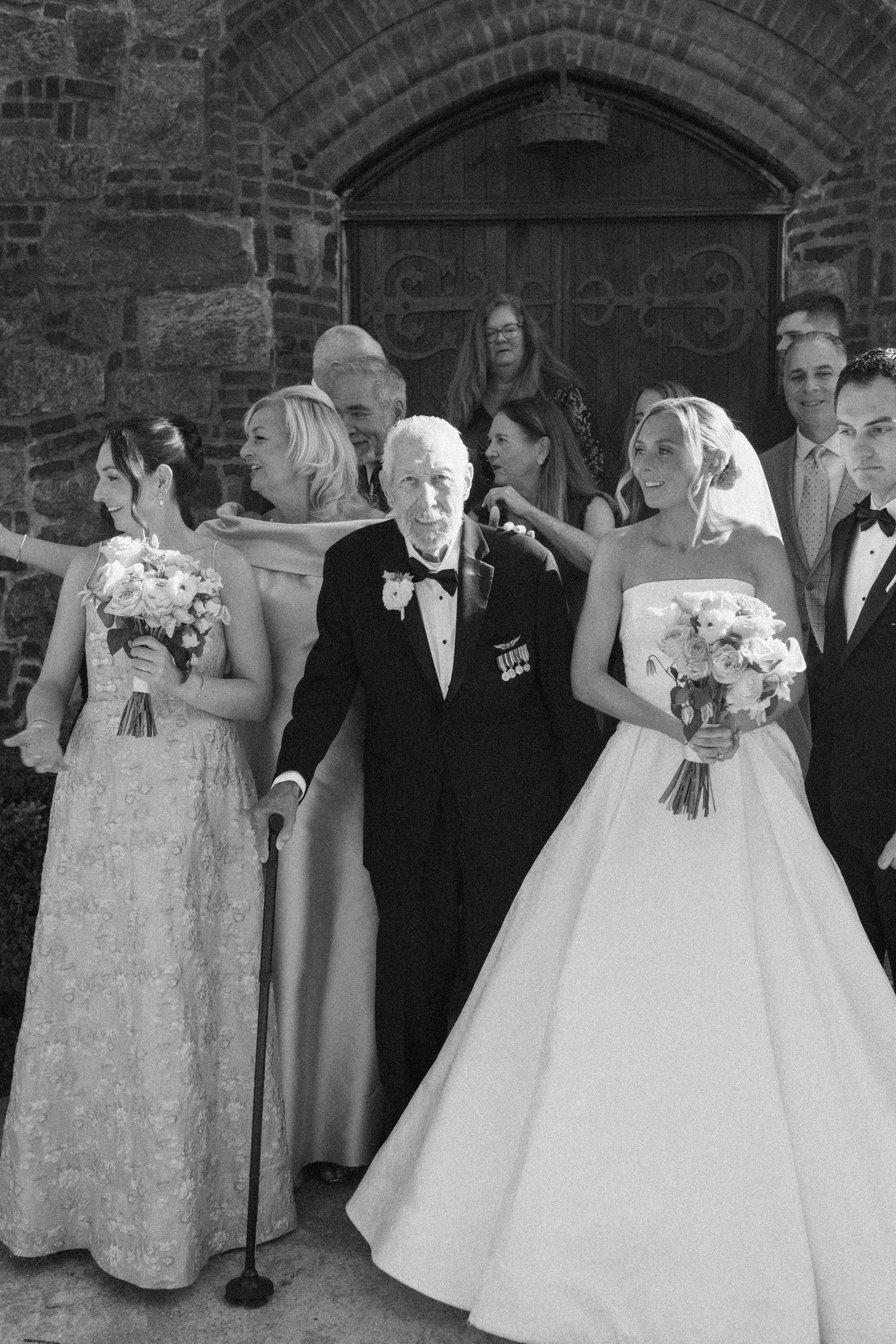 Black and white wedding photo of bride in ball gown with elderly veteran and bridal party outside stone church.