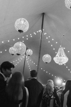 Black and white photo of elegant crystal chandeliers and string lights inside a wedding reception tent with guests.