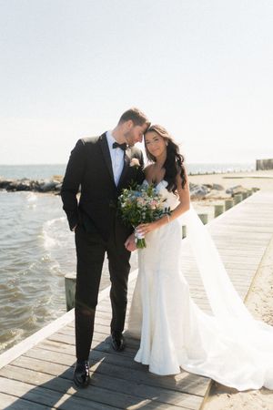 Bride and groom share a romantic moment on a waterfront pier, she holds a floral bouquet in her white gown.