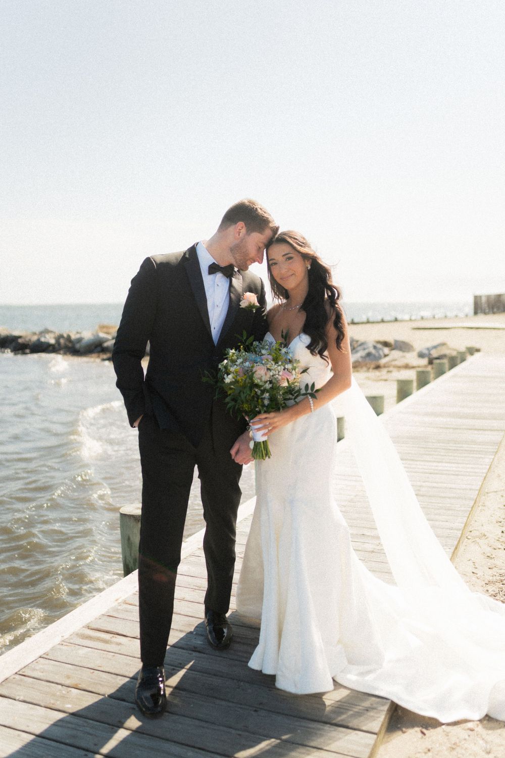 Bride and groom share a romantic moment on a waterfront pier, she holds a floral bouquet in her white gown.