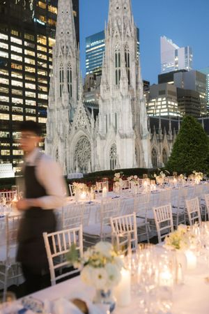 Elegant rooftop wedding reception with white chairs and candles, St. Patrick's Cathedral backdrop, NYC skyline at dusk.
