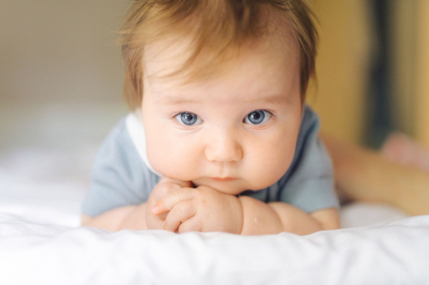 Adorable blue-eyed baby lying on white bed, resting chin on hands, wearing light blue onesie.