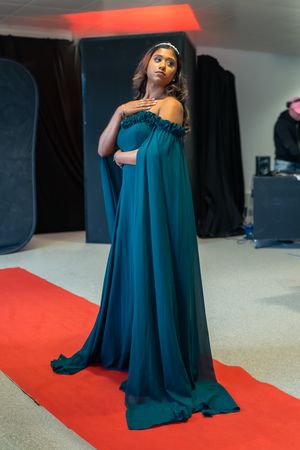 Pregnant woman in elegant teal off-shoulder gown with cape train poses on red carpet in photography studio.