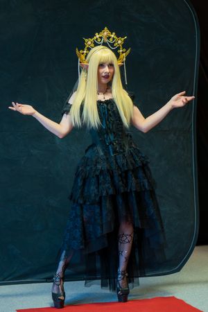Woman in gothic black tiered dress and gold crown poses with arms outstretched at cosplay event.