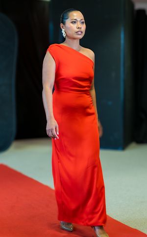 Woman in elegant red one-shoulder floor-length gown walking red carpet with pearl earrings and nude heels.
