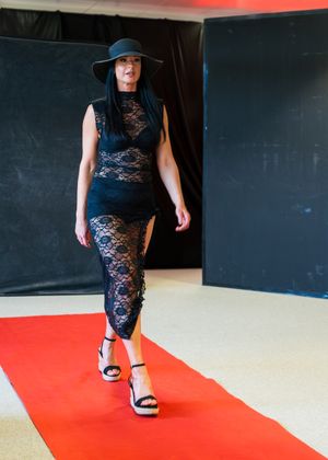 Woman in black lace dress and wide-brim hat walks red carpet runway at fashion show.