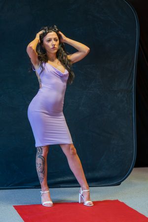 Woman in lavender bodycon dress and white heels poses on red carpet with tattoo visible on leg.