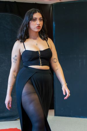 Young woman in black crop top and slit maxi skirt with tattoos poses confidently indoors.