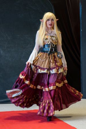 Cosplayer in elaborate burgundy and gold fantasy elf costume with blonde wig posing on red carpet.