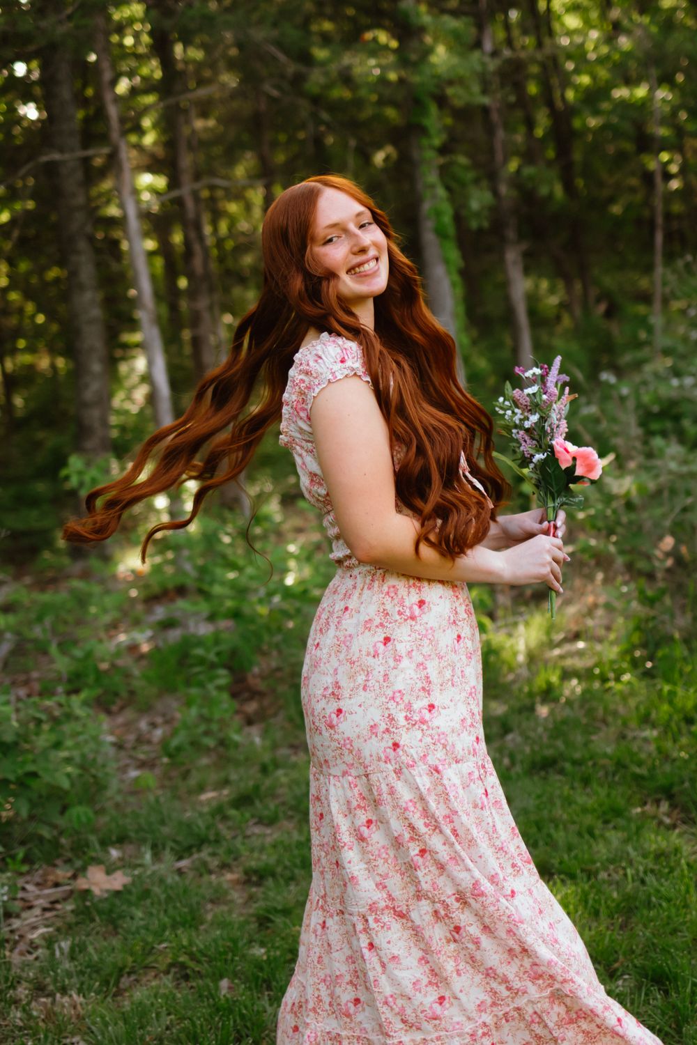 Smiling redhead woman in floral maxi dress holding flowers, twirling in a lush green forest setting.