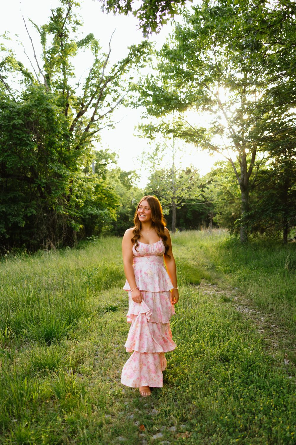 Young woman in pink floral tiered maxi dress walking barefoot on a sunlit forest path surrounded by lush green trees.