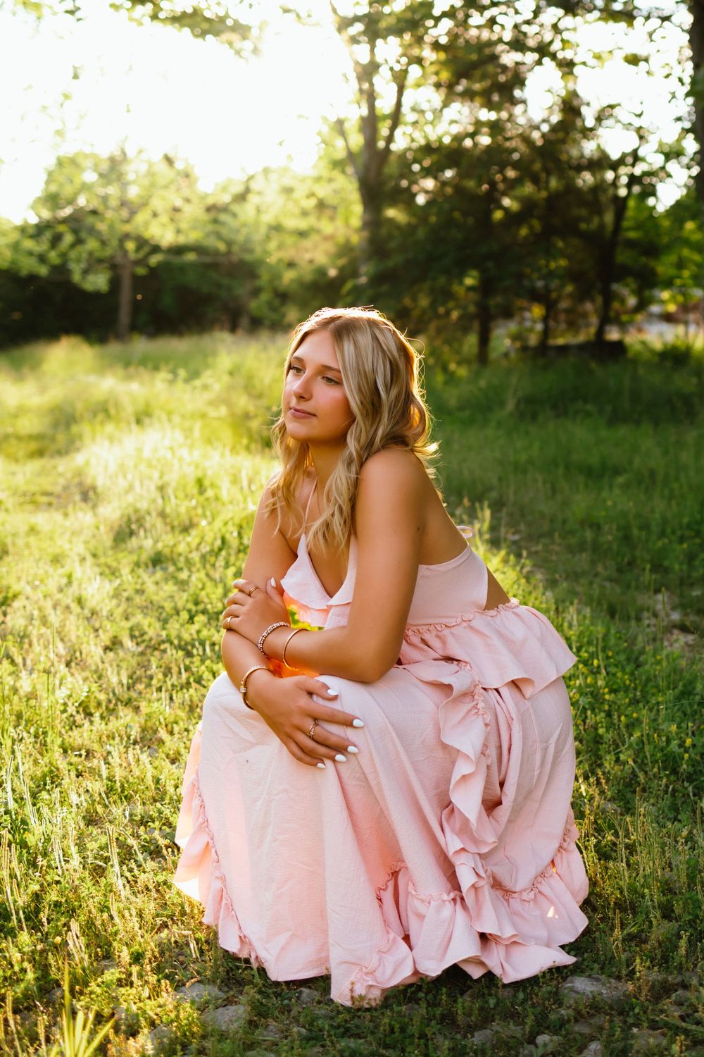 Blonde woman in pink ruffled dress sitting gracefully on green grass in golden sunlight outdoors.