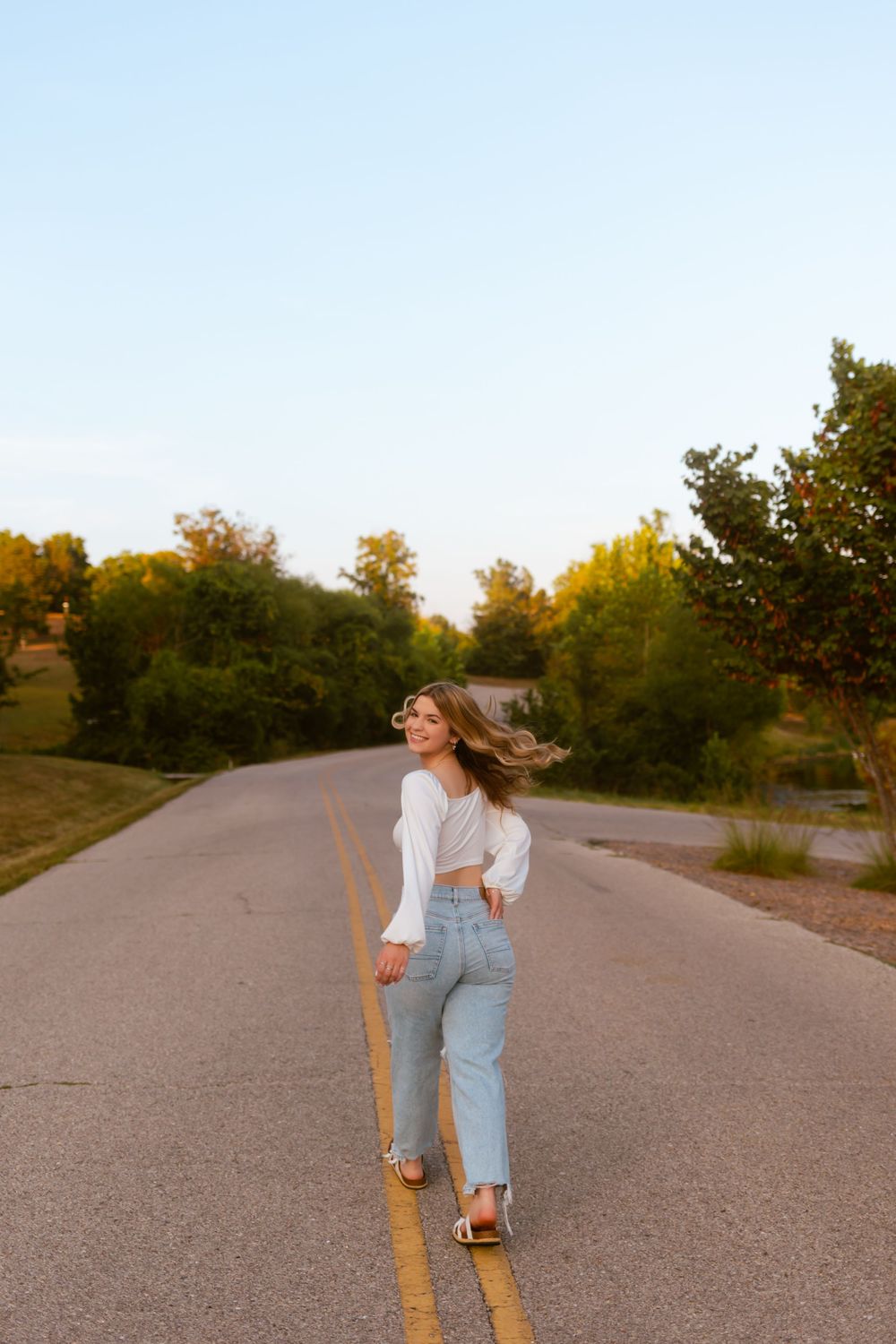 A person in light blue overalls and white top walks down a winding rural road during golden hour.