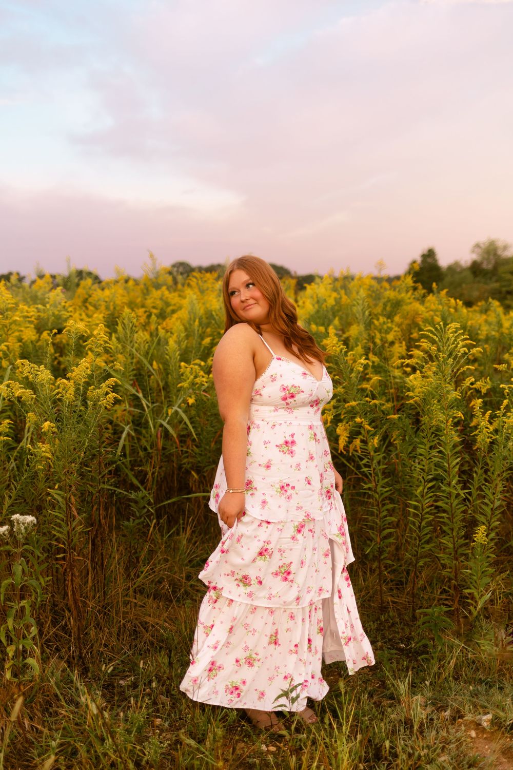 A woman in a white floral dress poses gracefully in a field of tall yellow wildflowers at sunset.