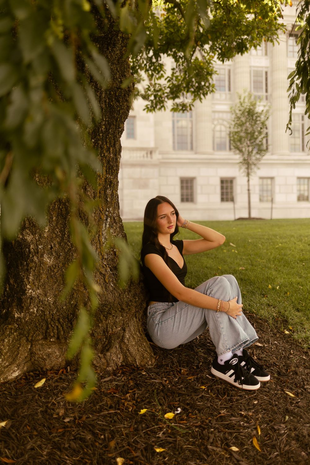 A person in casual attire relaxes under a shady tree on a grassy lawn near a grand building.