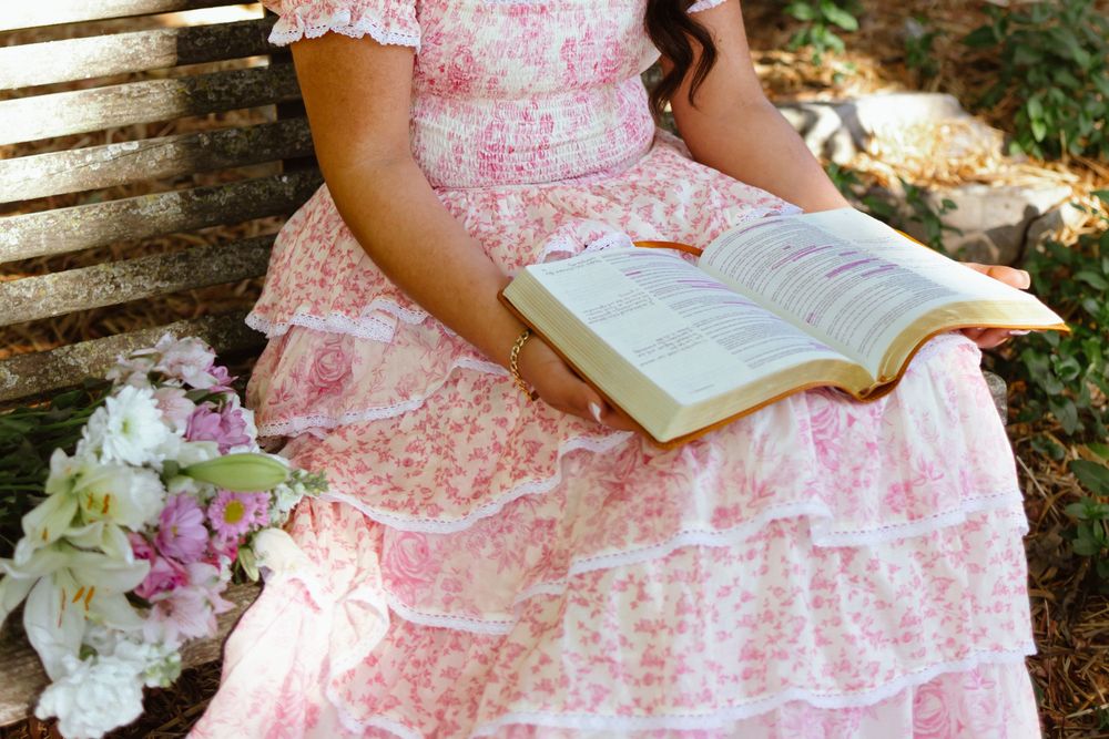 Woman in pink floral dress sitting on bench holding open Bible with bouquet of flowers beside her.
