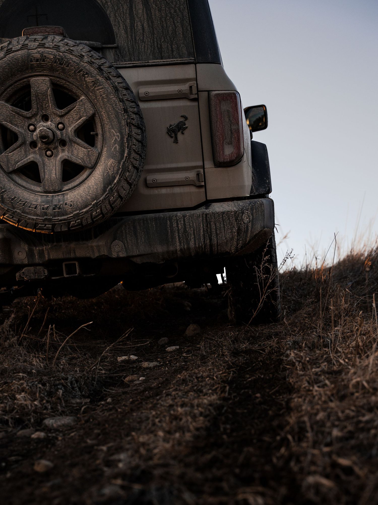 Muddy Ford Bronco rear view on rugged off-road dirt trail at dusk with spare tire mount visible.
