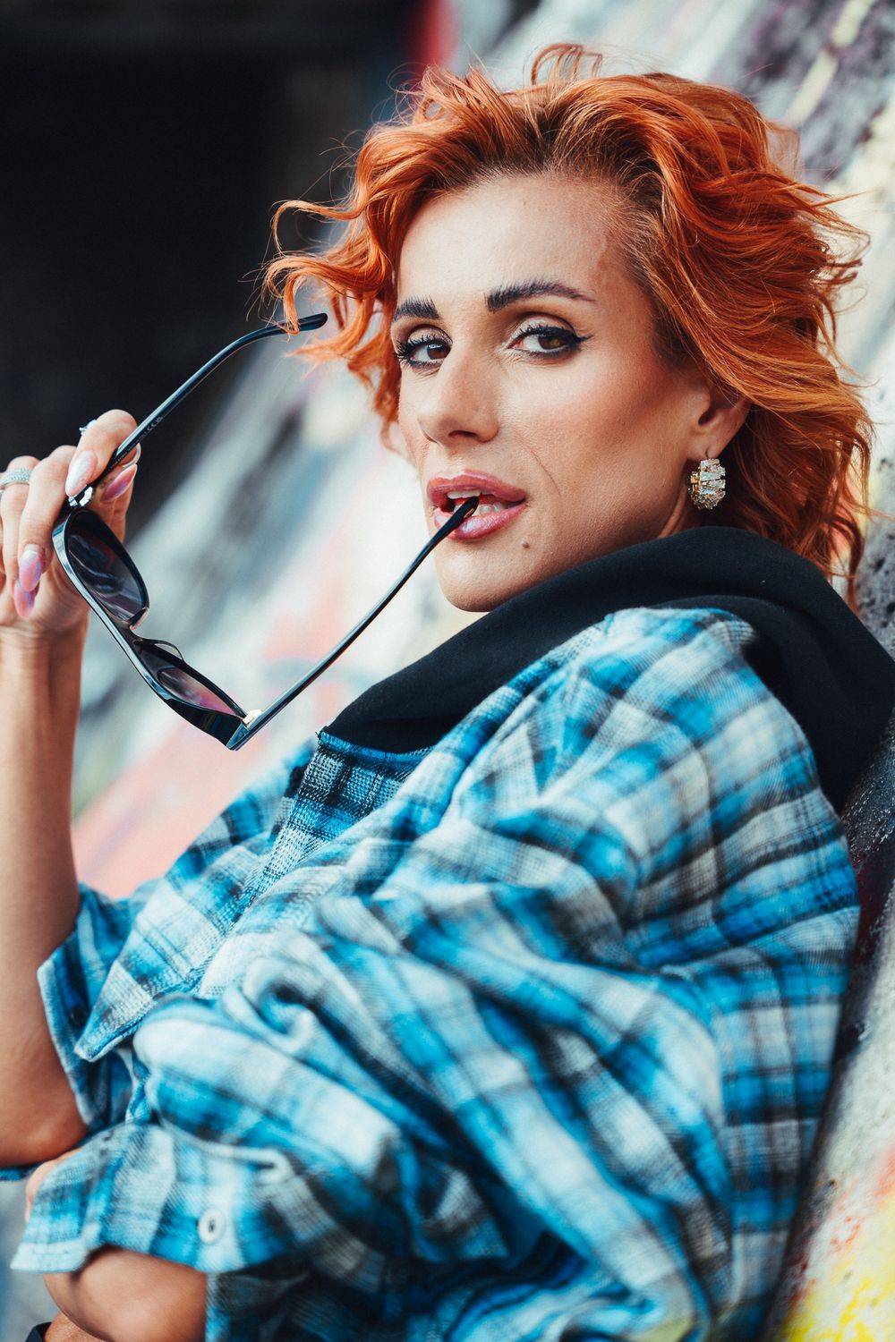 Red-haired woman in plaid shirt holding sunglasses near her mouth, posing against a graffiti wall.