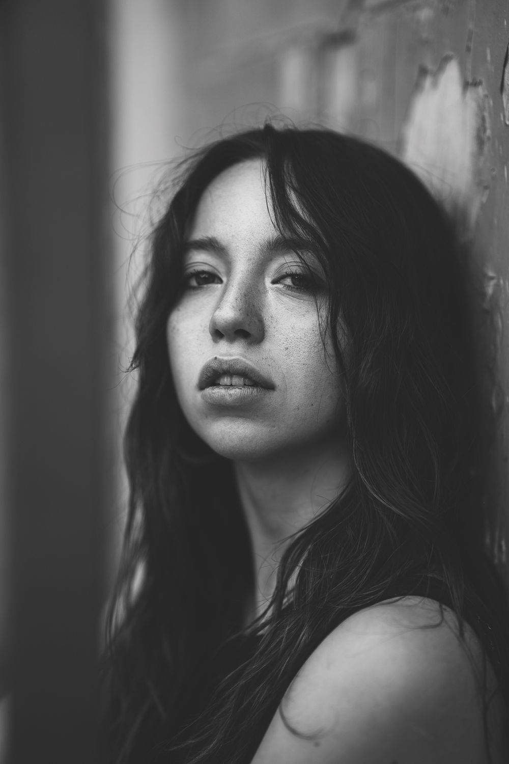 Black and white portrait of a young woman with long dark hair and freckles leaning against a wall.