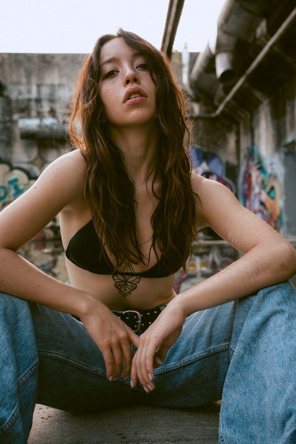 Young woman in black crop top and wide-leg jeans poses in urban setting with graffiti wall backdrop.
