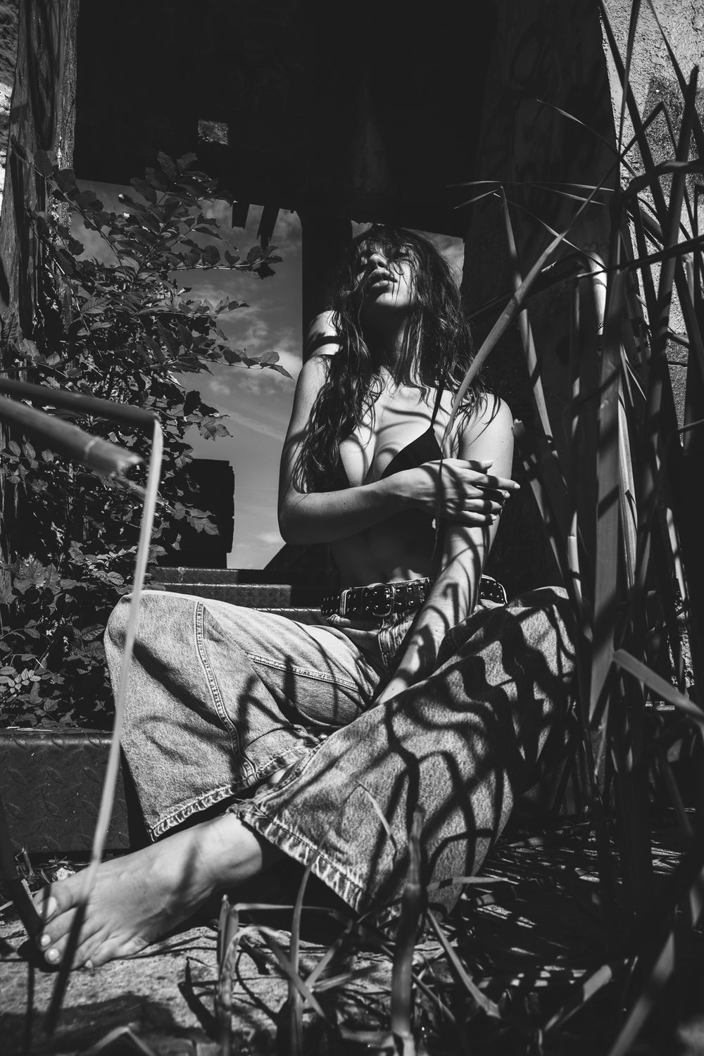 Black and white photo of a woman in bohemian skirt sitting on steps, surrounded by plants in dappled sunlight.