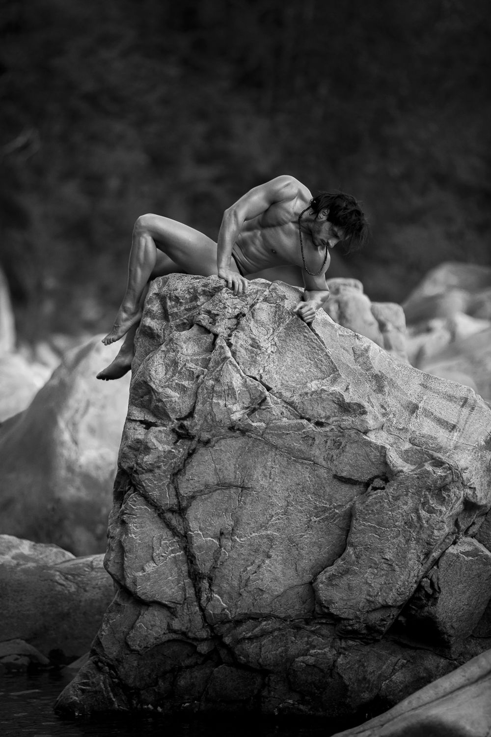 Black and white photo of a muscular nude man curled up on a large rock outdoors.