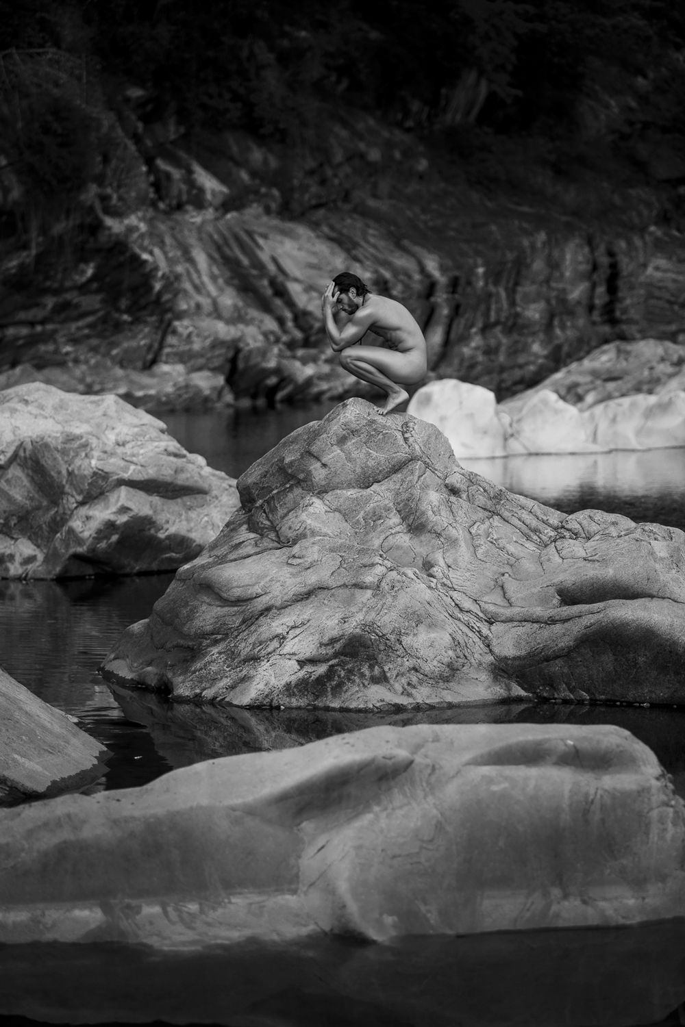 Black and white artistic photo of a nude figure crouching on a rock surrounded by water.