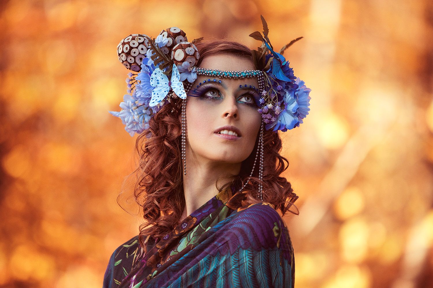 Beautiful woman with curly red hair wearing an ornate floral headdress with blue flowers against warm autumn bokeh.