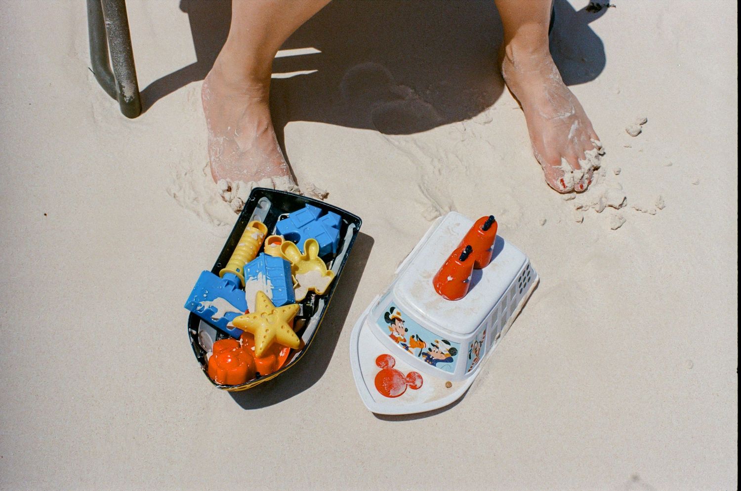 Person's feet on sandy beach beside colorful toy-filled sandal and Mickey Mouse boat sandal.