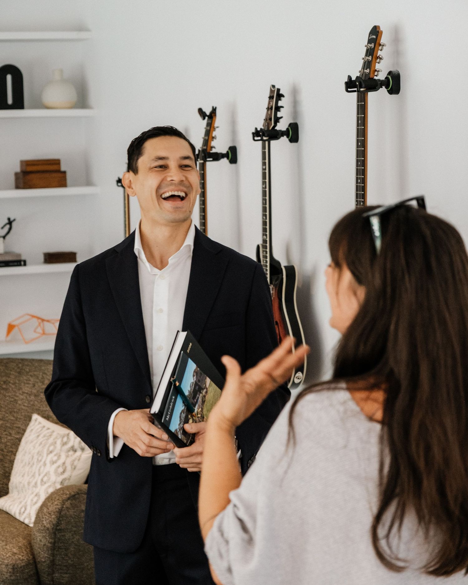 Man in black blazer laughing while holding books, with guitars on wall mount in bright modern office.