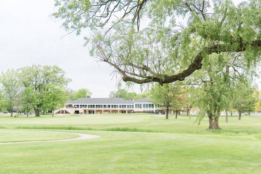 Lush green golf course with a large clubhouse building framed by weeping willow trees on a cloudy day.
