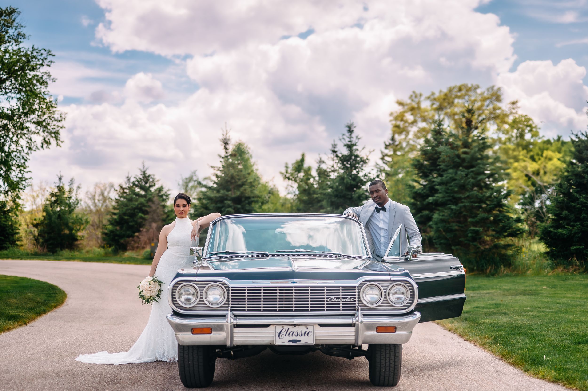 Bride and groom posing with a classic vintage car on a scenic driveway surrounded by lush green trees.