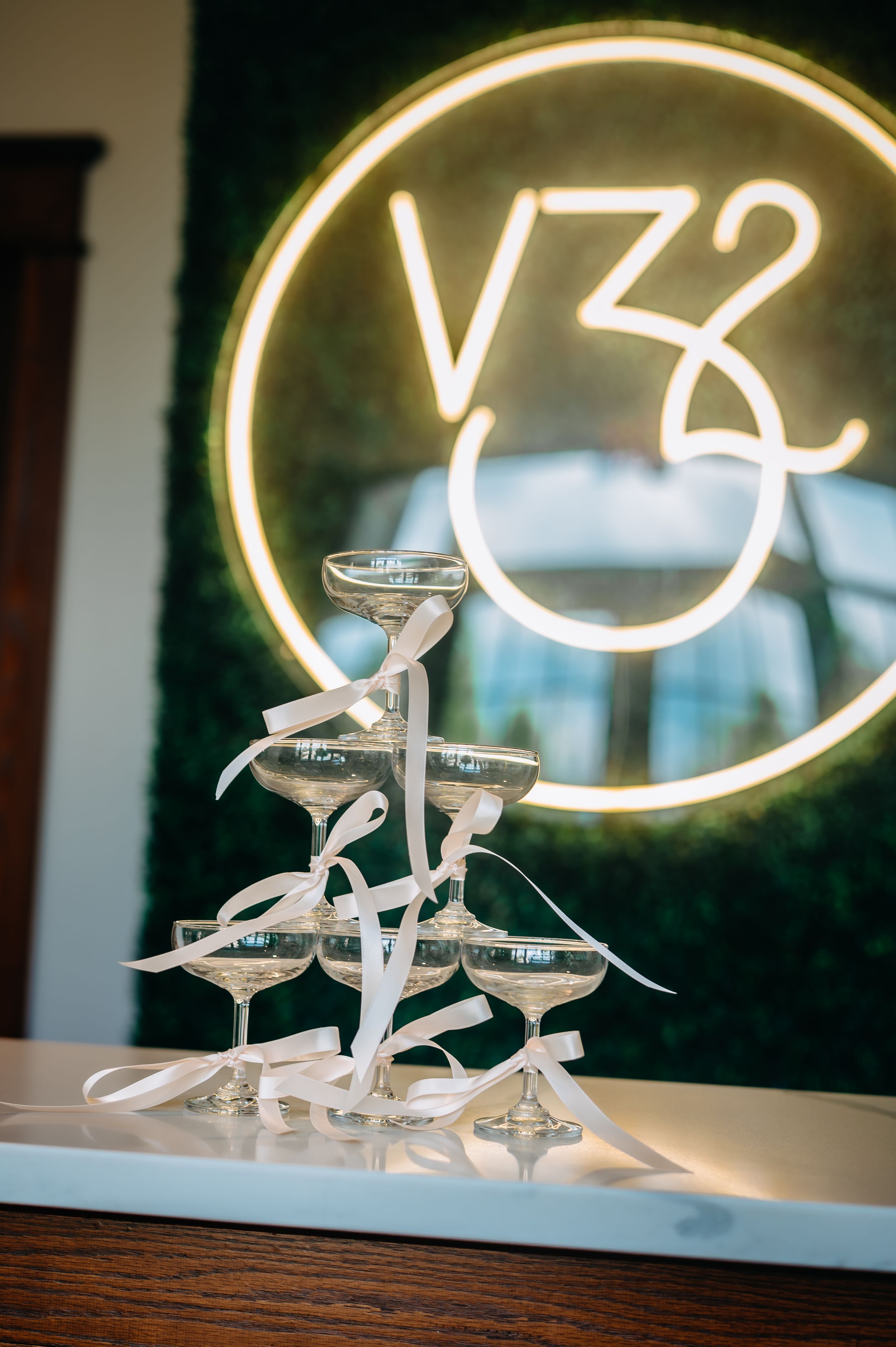 Champagne tower with white ribbons in front of a glowing V32 neon sign on a green moss wall.