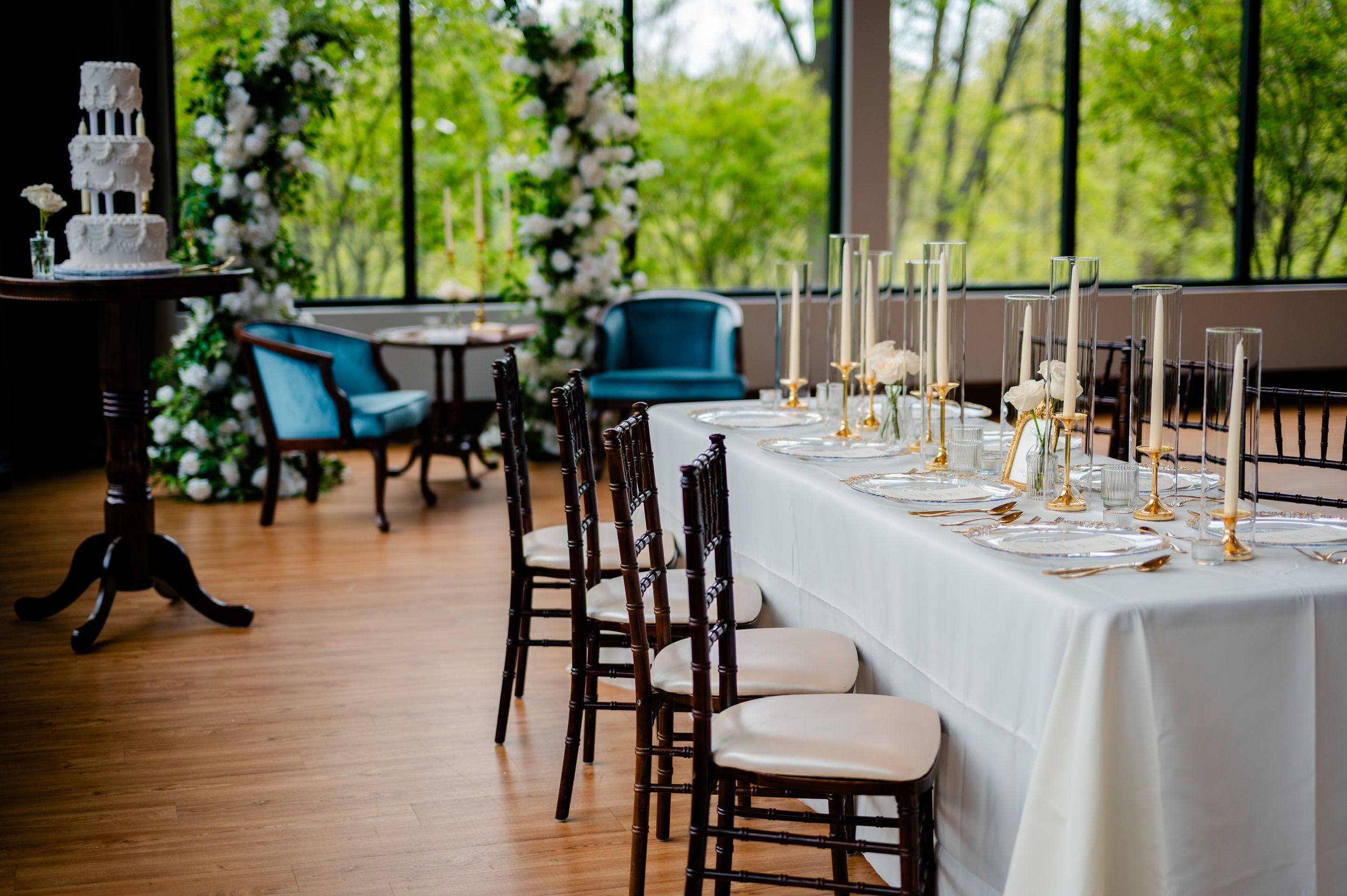 Elegant wedding reception setup with white tablecloths, gold candles, chiavari chairs, and floral arch near large windows.